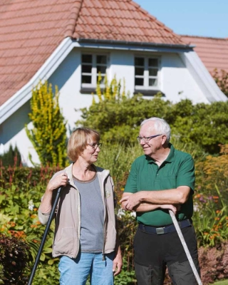 Ein aelteres Paar steht im Vorgarten ihrer Immobilie.
